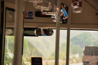 The interior of a bus is visible, focusing on the driver's area. A driver in a blue shirt is seated, visible through a rearview mirror. Some informational signs and a digital clock displaying time are mounted near the ceiling. The bus windows provide a view to the outside where greenery and a blurred vehicle can be seen.