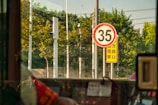A speed limit sign displaying the number 35 is prominently visible, with a backdrop of lush green trees. Adjacent to it is a yellow sign with text in a foreign script. The view appears to be from inside a vehicle, with parts of the interior and a window frame visible.