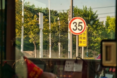 A speed limit sign displaying the number 35 is prominently visible, with a backdrop of lush green trees. Adjacent to it is a yellow sign with text in a foreign script. The view appears to be from inside a vehicle, with parts of the interior and a window frame visible.