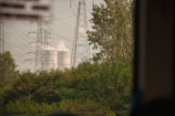 Tall electricity pylons and transmission lines dominate the landscape, standing amidst lush green foliage. In the background, cooling towers are visible, emitting steam or smoke into the air.