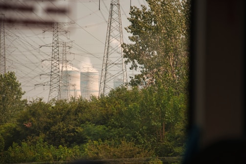 Tall electricity pylons and transmission lines dominate the landscape, standing amidst lush green foliage. In the background, cooling towers are visible, emitting steam or smoke into the air.