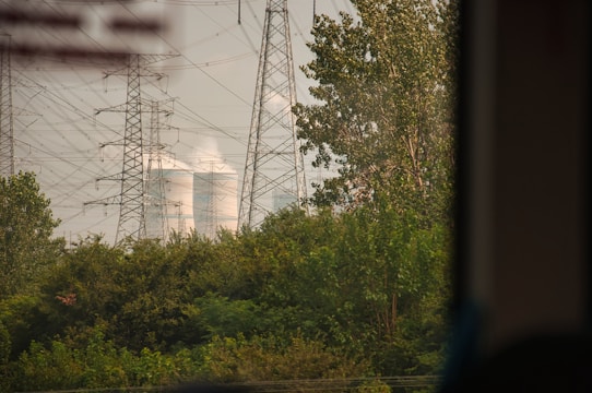 Tall electricity pylons and transmission lines dominate the landscape, standing amidst lush green foliage. In the background, cooling towers are visible, emitting steam or smoke into the air.