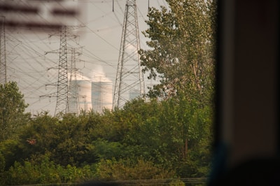 Tall electricity pylons and transmission lines dominate the landscape, standing amidst lush green foliage. In the background, cooling towers are visible, emitting steam or smoke into the air.