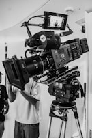 A professional film camera setup on a tripod, featuring a large lens and a mounted monitor displaying an image of a person. The camera has multiple controls and is positioned indoors with part of a person visible in the background.