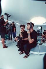 A group of people is engaged in a video production setting. Two individuals in the foreground are seated; one of them is wearing a headset and holding a device with various buttons, the other appears to be observing. In the background, three other people are standing, with one person looking at a video camera setup on a tripod. The room has futuristic, minimalist decor with white walls and a large, circular light fixture on the ceiling.