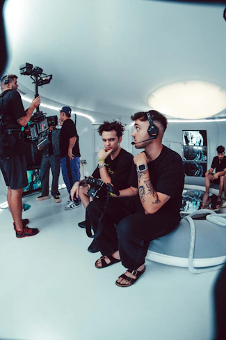 A group of people is engaged in a video production setting. Two individuals in the foreground are seated; one of them is wearing a headset and holding a device with various buttons, the other appears to be observing. In the background, three other people are standing, with one person looking at a video camera setup on a tripod. The room has futuristic, minimalist decor with white walls and a large, circular light fixture on the ceiling.