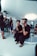 A group of people is engaged in a video production setting. Two individuals in the foreground are seated; one of them is wearing a headset and holding a device with various buttons, the other appears to be observing. In the background, three other people are standing, with one person looking at a video camera setup on a tripod. The room has futuristic, minimalist decor with white walls and a large, circular light fixture on the ceiling.