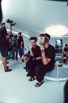 A group of people is engaged in a video production setting. Two individuals in the foreground are seated; one of them is wearing a headset and holding a device with various buttons, the other appears to be observing. In the background, three other people are standing, with one person looking at a video camera setup on a tripod. The room has futuristic, minimalist decor with white walls and a large, circular light fixture on the ceiling.