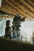 A person operates a professional camera on a tripod indoors, in a room with rustic wooden beams on the ceiling and a soft natural light filtering through the window.