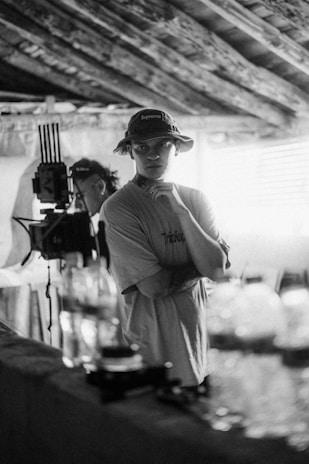A black and white photograph of a person wearing a hat and t-shirt, standing indoors near film equipment. The person appears to be thoughtful, with a hand resting on their chin. The setting has a rustic, textured ceiling and there are out-of-focus bottles and containers in the foreground.
