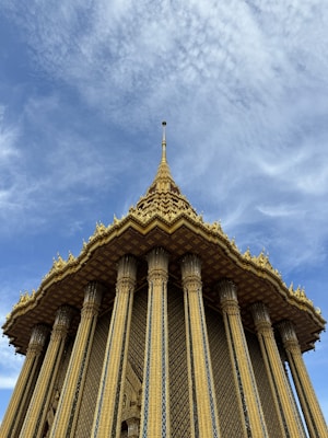 A majestic golden temple with intricate patterns and tall, ornate columns reaching up towards a partly cloudy sky. The roof is richly decorated with elaborate spires and detailed embellishments, creating a stunning contrast with the blue sky.