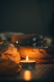 A small tealight candle casts a warm glow, surrounded by a knitted blanket and a blurred dark backdrop. The lighting creates a cozy and intimate atmosphere, with soft shadows and highlights on the textured fabric.