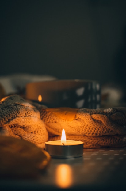 A small tealight candle casts a warm glow, surrounded by a knitted blanket and a blurred dark backdrop. The lighting creates a cozy and intimate atmosphere, with soft shadows and highlights on the textured fabric.