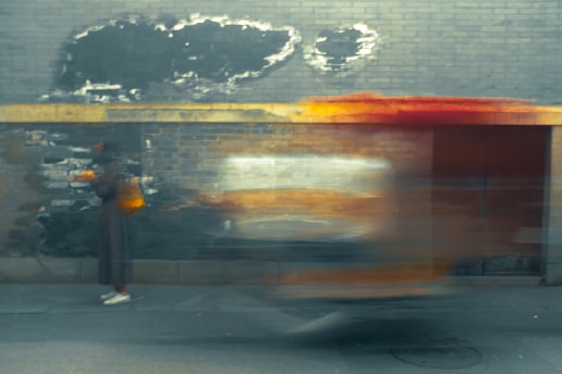 A blurry scene depicting urban motion, with a person dressed in dark clothing and white shoes standing against a worn, gray brick wall. The wall shows areas of peeling paint and weathering. A brightly colored object, possibly a moving vehicle, creates a streak of motion in front of the person, suggesting speed and movement.