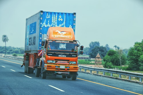 An orange Isuzu truck with a large container on its back is driving on a highway. The truck has signage indicating 'K.B Logistics' and contains decorative elements on the front. The container is mostly blue with white text and appears to be used for transporting goods. The highway is surrounded by greenery, with trees and bushes visible in the background. It's a bright day with clear visibility.