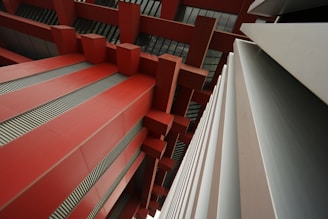 An abstract architectural view featuring large red beams and geometric structures. Strong lines and shapes create a modern, industrial aesthetic, with a focus on symmetry and perspective.