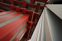 An abstract architectural view featuring large red beams and geometric structures. Strong lines and shapes create a modern, industrial aesthetic, with a focus on symmetry and perspective.