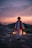 A man wearing traditional attire with a red turban and scarf stands outdoors on a rocky surface at sunset, holding two lanterns that emit a warm glow. The sky above is filled with clouds tinged by the fading sunlight, creating a tranquil scene.