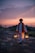 A man wearing traditional attire with a red turban and scarf stands outdoors on a rocky surface at sunset, holding two lanterns that emit a warm glow. The sky above is filled with clouds tinged by the fading sunlight, creating a tranquil scene.