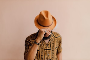 A person wearing a patterned shirt and an orange-brown hat is slightly tilting their head downward while holding the brim of the hat with one hand. The background is plain and beige, enhancing the focus on the individual's attire.