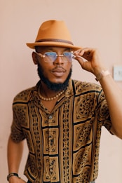 A stylish person wearing a patterned shirt and a tan fedora hat is adjusting their sunglasses. They have a well-groomed beard and are accessorized with a beaded necklace and a watch.