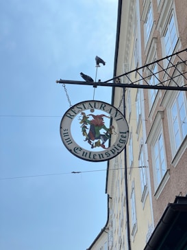 A metal sign hangs from an ornate bracket attached to the side of a building. The round sign features the words 'Restaurant zum Eulenspiegel' around a colorful illustration of a jester. Two birds are perched on top of the sign against a clear blue sky, and the side of the building with multiple windows is visible.