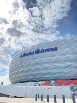 A large, modern stadium with a unique, textured exterior featuring the name 'Allianz Arena' in bold, blue letters. The sky above is filled with scattered, fluffy clouds, creating a bright and open atmosphere. In front of the stadium, there is a mural or advertisement showing a football tournament with vibrant red colors.