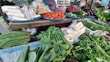 A vibrant market stall displays an assortment of fresh vegetables and packaged food items. Tomatoes, cucumbers, eggplants, and chillies are neatly arranged on blue trays. Various leafy greens and bundled herbs are piled in front, alongside packaged corn. The colorful produce is set against a rustic wooden table.