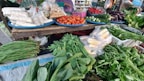 A vibrant market stall displays an assortment of fresh vegetables and packaged food items. Tomatoes, cucumbers, eggplants, and chillies are neatly arranged on blue trays. Various leafy greens and bundled herbs are piled in front, alongside packaged corn. The colorful produce is set against a rustic wooden table.