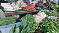 A vibrant market stall displays an assortment of fresh vegetables and packaged food items. Tomatoes, cucumbers, eggplants, and chillies are neatly arranged on blue trays. Various leafy greens and bundled herbs are piled in front, alongside packaged corn. The colorful produce is set against a rustic wooden table.