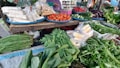A vibrant market stall displays an assortment of fresh vegetables and packaged food items. Tomatoes, cucumbers, eggplants, and chillies are neatly arranged on blue trays. Various leafy greens and bundled herbs are piled in front, alongside packaged corn. The colorful produce is set against a rustic wooden table.