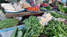 A vibrant market stall displays an assortment of fresh vegetables and packaged food items. Tomatoes, cucumbers, eggplants, and chillies are neatly arranged on blue trays. Various leafy greens and bundled herbs are piled in front, alongside packaged corn. The colorful produce is set against a rustic wooden table.