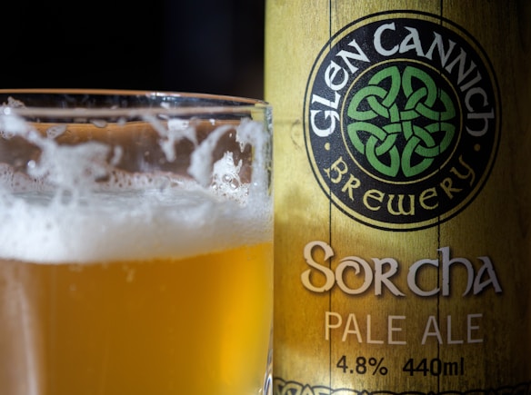 A close-up shot of a pale ale beer with foam in a glass alongside a can. The can bears the logo and name of Glen Cannich Brewery and denotes the specific beer as Sorcha Pale Ale with an alcohol content of 4.8% and a volume of 440ml. The design features Celtic-inspired artwork and text.