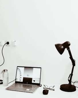 A minimalist workspace setup featuring a laptop displaying black and white photography on its screen. Next to it is a water bottle with a metallic lid, an ergonomic computer mouse, and a pair of eyeglasses. A black desk lamp with an adjustable arm is positioned to the right, casting a subtle shadow on the white surface.