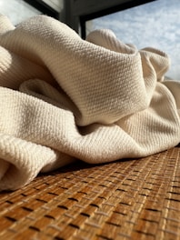 Soft, cream-colored fabric with a textured surface is draped casually over a woven, wooden-textured surface. Sunlight filters through a window in the background, casting light and shadow on the scene.