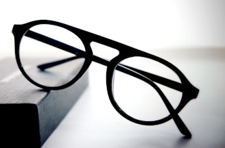 A pair of round black eyeglasses with a thin frame is placed on a dark rectangular object with a soft gradient background. The glasses appear to be stylish and simple, with clear lenses and bold frames.