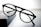 A pair of round black eyeglasses with a thin frame is placed on a dark rectangular object with a soft gradient background. The glasses appear to be stylish and simple, with clear lenses and bold frames.