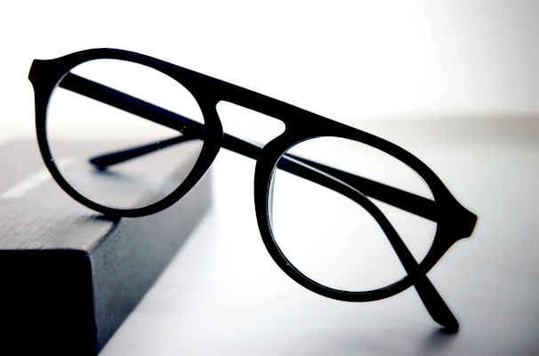 A pair of round black eyeglasses with a thin frame is placed on a dark rectangular object with a soft gradient background. The glasses appear to be stylish and simple, with clear lenses and bold frames.