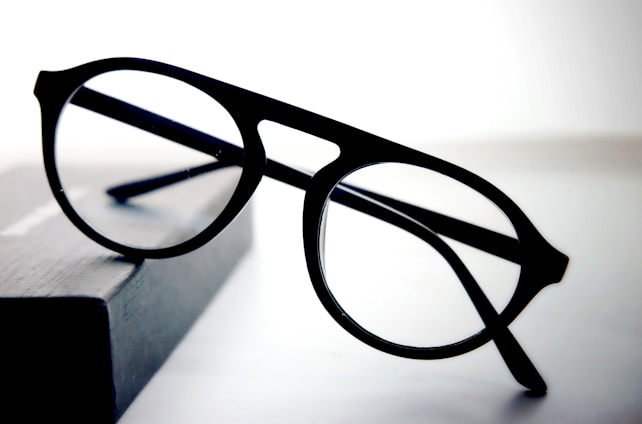A pair of round black eyeglasses with a thin frame is placed on a dark rectangular object with a soft gradient background. The glasses appear to be stylish and simple, with clear lenses and bold frames.
