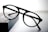 A pair of round black eyeglasses with a thin frame is placed on a dark rectangular object with a soft gradient background. The glasses appear to be stylish and simple, with clear lenses and bold frames.