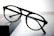 A pair of round black eyeglasses with a thin frame is placed on a dark rectangular object with a soft gradient background. The glasses appear to be stylish and simple, with clear lenses and bold frames.