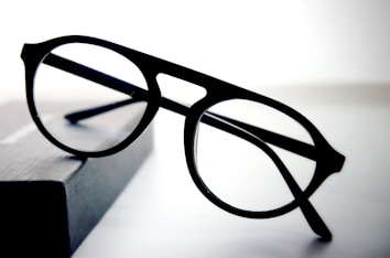 A pair of round black eyeglasses with a thin frame is placed on a dark rectangular object with a soft gradient background. The glasses appear to be stylish and simple, with clear lenses and bold frames.