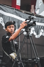 A young man operates a professional video camera on a tripod, wearing headphones and an equipment vest. He appears focused on his task. The background features a black and white fabric banner and a building with red and white colors.