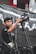 A young man operates a professional video camera on a tripod, wearing headphones and an equipment vest. He appears focused on his task. The background features a black and white fabric banner and a building with red and white colors.