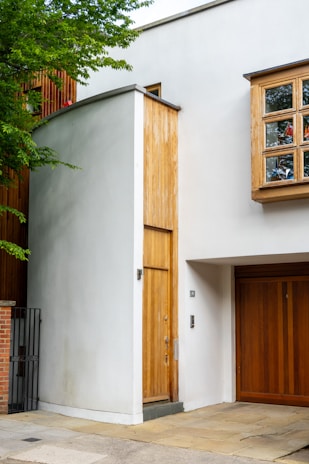 A modern building façade features clean lines and a minimalist design. A tall, narrow wooden door is set into a white wall, complemented by large wooden-framed windows. The entrance is adjacent to a garage door of similar wood. Lush green foliage partially frames the building.