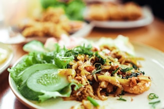 A plate of stir-fried noodles garnished with fresh herbs is accompanied by sliced cucumber and lettuce. The dish appears to have pieces of shrimp and is served on a ceramic plate with a blurred background suggesting more food.