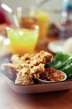 A plate with crispy fried chicken pieces accompanied by a serving of chili sauce and fresh green lettuce. In the background, there's a blurred glass of yellow-orange beverage with a straw, suggesting a refreshing drink.