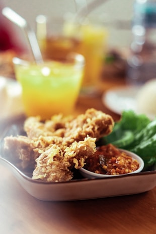 A plate with crispy fried chicken pieces accompanied by a serving of chili sauce and fresh green lettuce. In the background, there's a blurred glass of yellow-orange beverage with a straw, suggesting a refreshing drink.