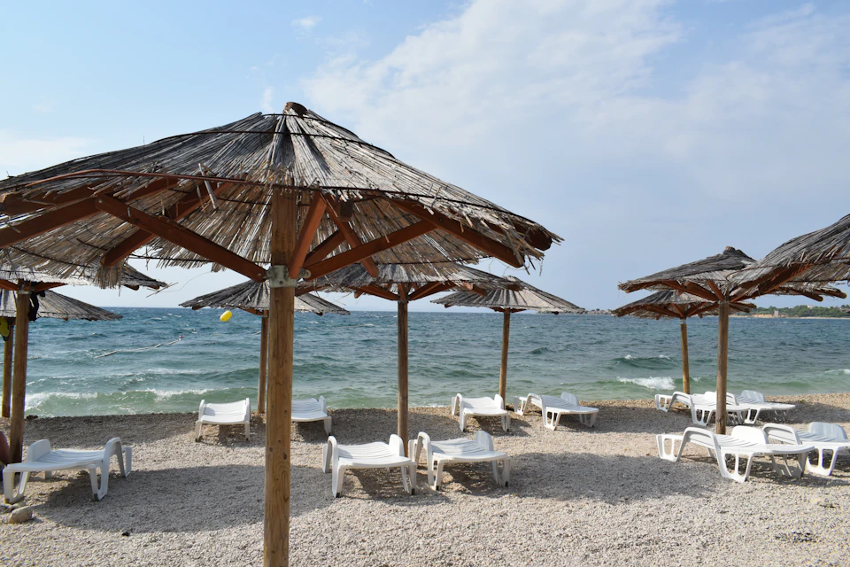 Strohschirme und weiße Liegen an einem Kiesstrand, bewegte See