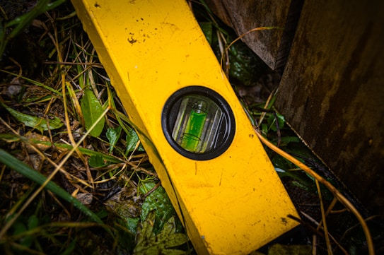 A bright yellow level tool lies on top of wet grass and foliage near a wooden structure. The circular vial in the center of the tool is visible and contrast sharply with the green leaves surrounding it. Moisture is evident on the leaves and tool, indicating a damp environment.